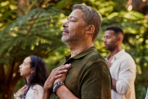 Group of multiethnic people standing outdoors with eyes closed and hands on their hearts during a meditation session. Multiracial adults practicing gratitude and mindfulness together. Mature latin man connecting to inner peace and presence at wellness retreat center in nature.