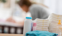 Close up of baby products sitting on a table in a baby's nursery. A mom is changing a baby's diaper in the background.